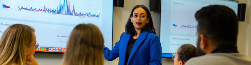 lecturer speaking to students with a white board displaying a graph in the back ground