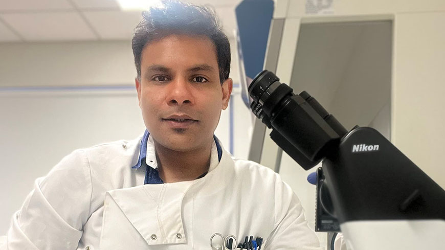 Dr Prashant Bharadwaj wears a white lab coat in the lab, there is a black microscope in the corner.