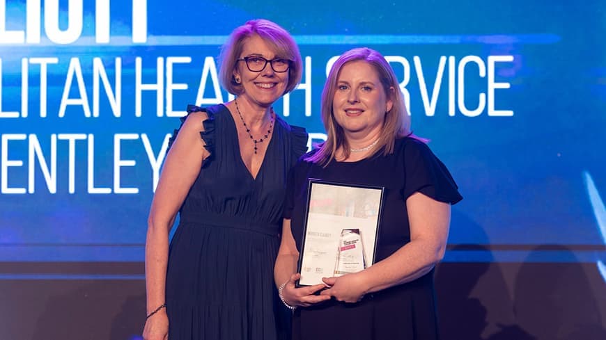 Professor Tracey Moroney OAM wears a dark blue frilled dress and glasses and stands with Noreen Elliott who wears a black dress and holds a framed award.