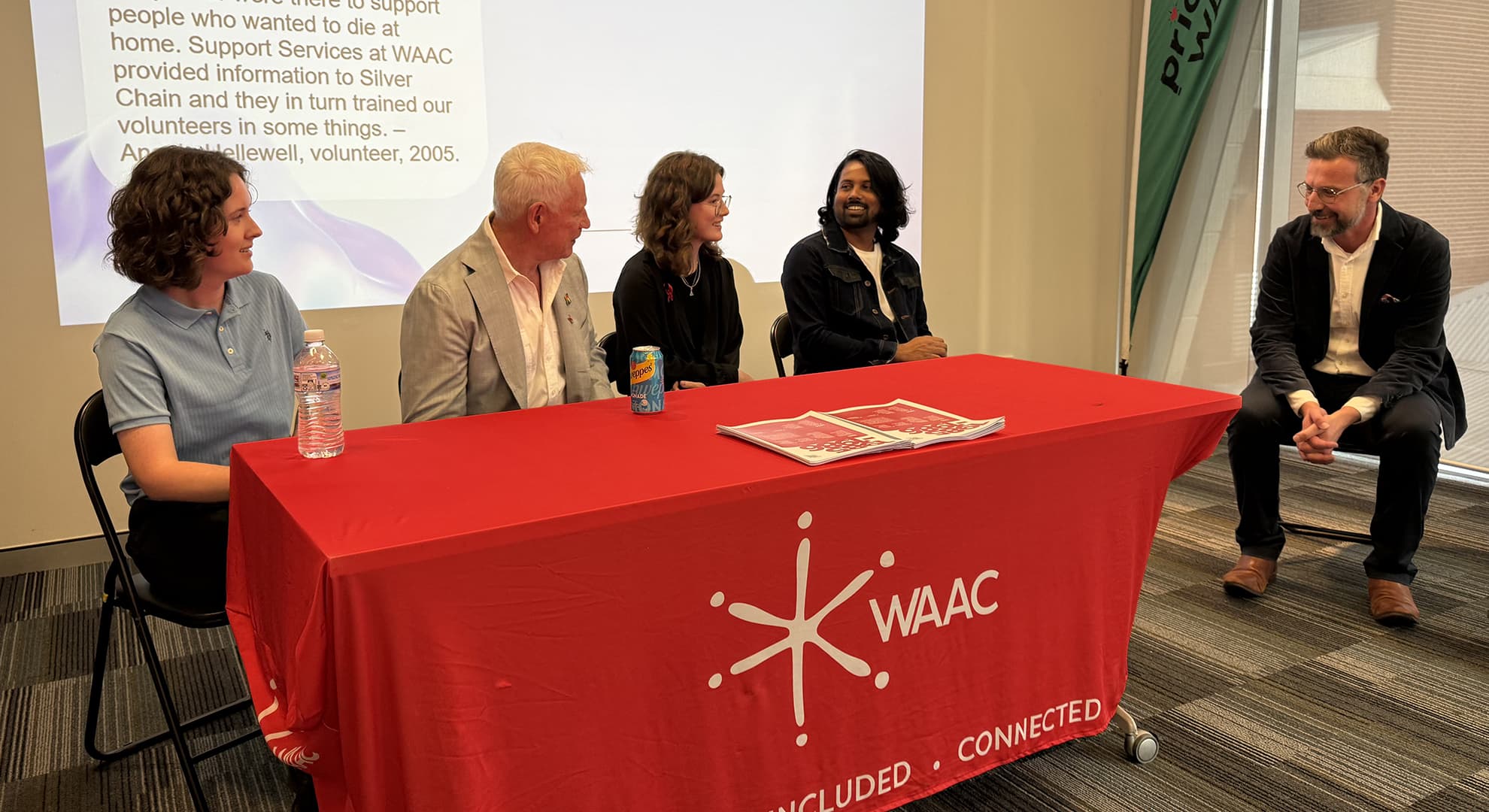 a panel of 4 people sitting behind a desk with a red tablecloth, another person sitting on the right