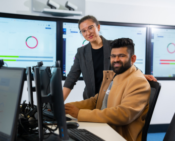 Two students in a computer lab, one seated at a workstation and the other standing beside them, both smiling. Large screens in the background display data visualizations.