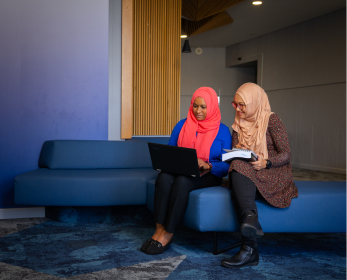 Two women sitting on a blue couch, one using a laptop while the other takes notes, collaborating in a modern study space.