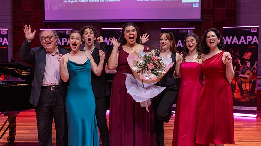 A pianist poses with WAAPA students on stage, they are all dressed formally and are grinning and excited.