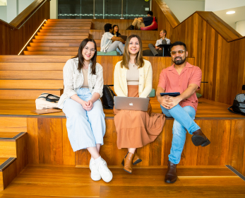 Three ECU students sitting on wooden tiered seating with laptops and notebooks in the Joondalup campus library.