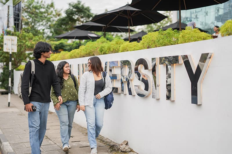 Three students walk and chat outside the university campus, set against a lush background of greenery and black umbrellas.