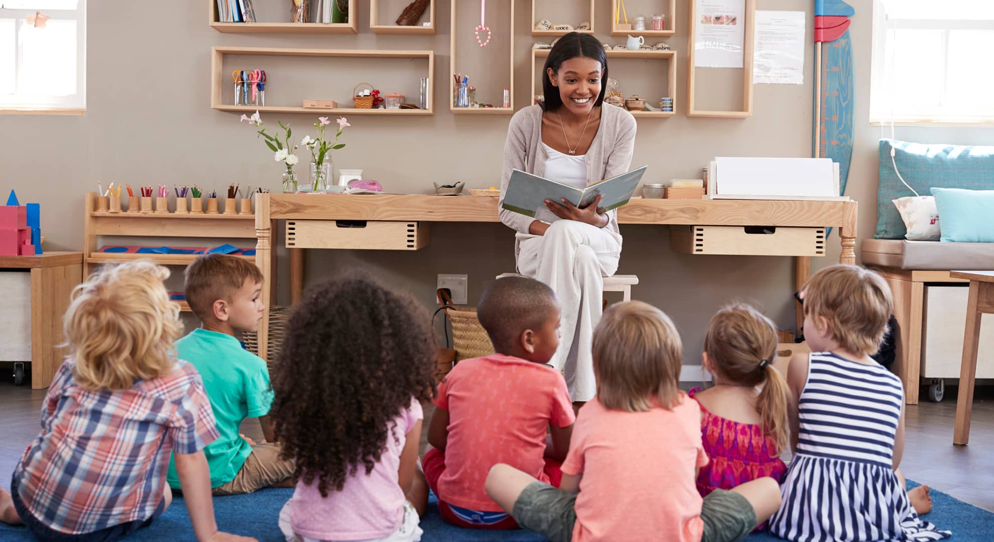 Classroom teacher smiling, sitting on chair and reading a story to a group of seven children. The children are facing the teacher, sitting on a mat, with their backs to the camera.