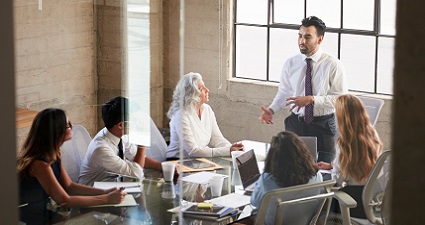Four people sitting down at a boardroom table, looking up at man presenting