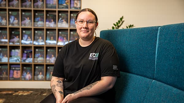 young woman wearing glasses is sitting on a couch with ECU course guides stacked on a display behind her.