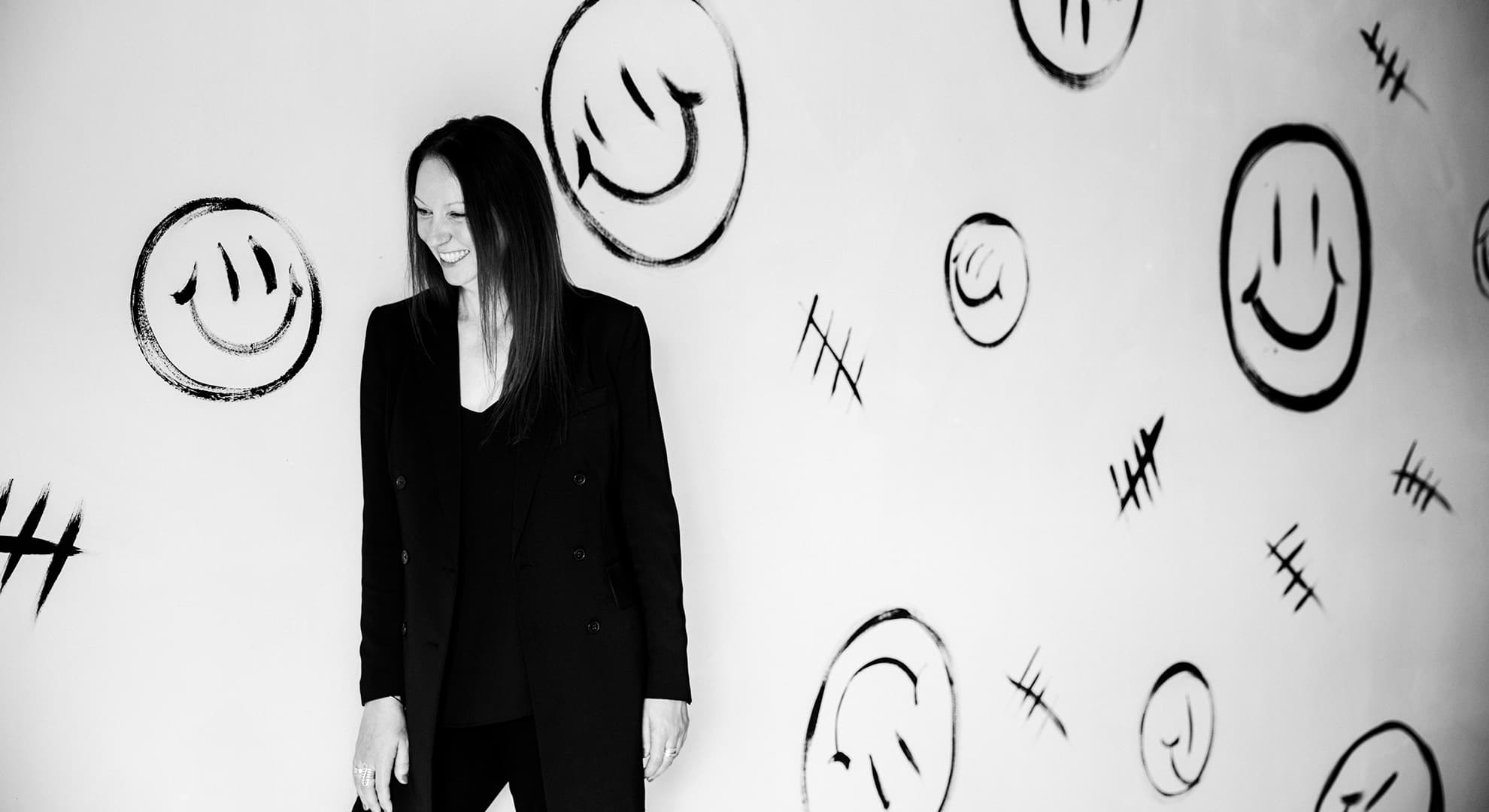 A black and white horizontal photo of Professor Narelle Lemon standing in a black suit, smiling, looking away from camera. Behind her is an artistic backdrop of smiley faces.