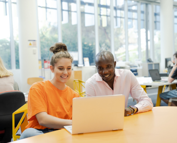 Two ECU students seated at a brightly lit library table, smiling and engaged as they look at a laptop together. Large windows and study spaces in the background suggest a welcoming campus environment during the two-week Orientation program.