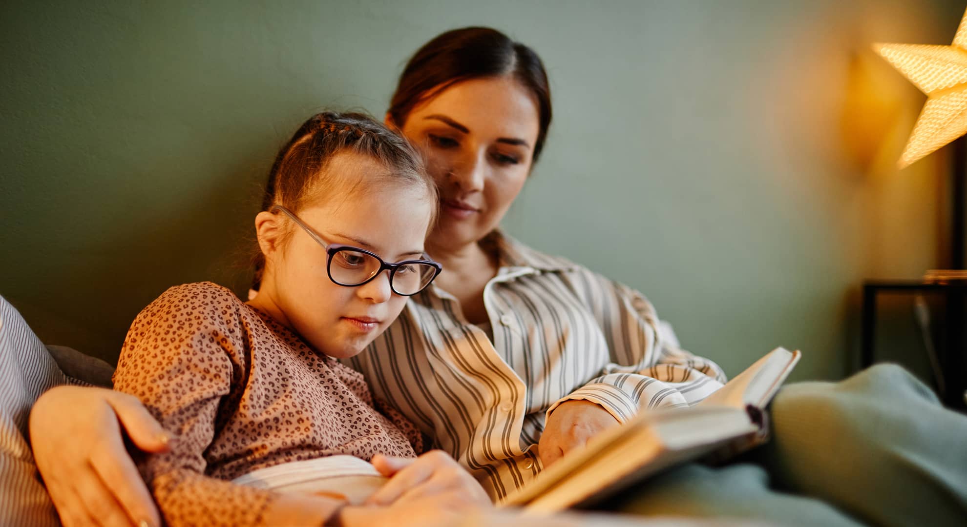 Mother and daughter with down syndrome rugged up in bed together reading a children's book.