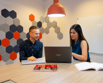 Two people seated at a meeting table looking at a laptop screen together in a bright study room with books and notebooks on the table.