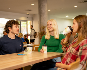 Three students sit together at a wooden table indoors, talking and smiling while holding takeaway coffee cups.