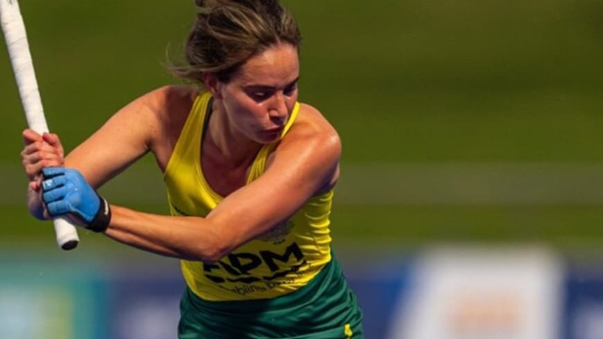 hockey sportswoman in yellow singlet, green bottoms swings white hockey stick above her head blurred green in background