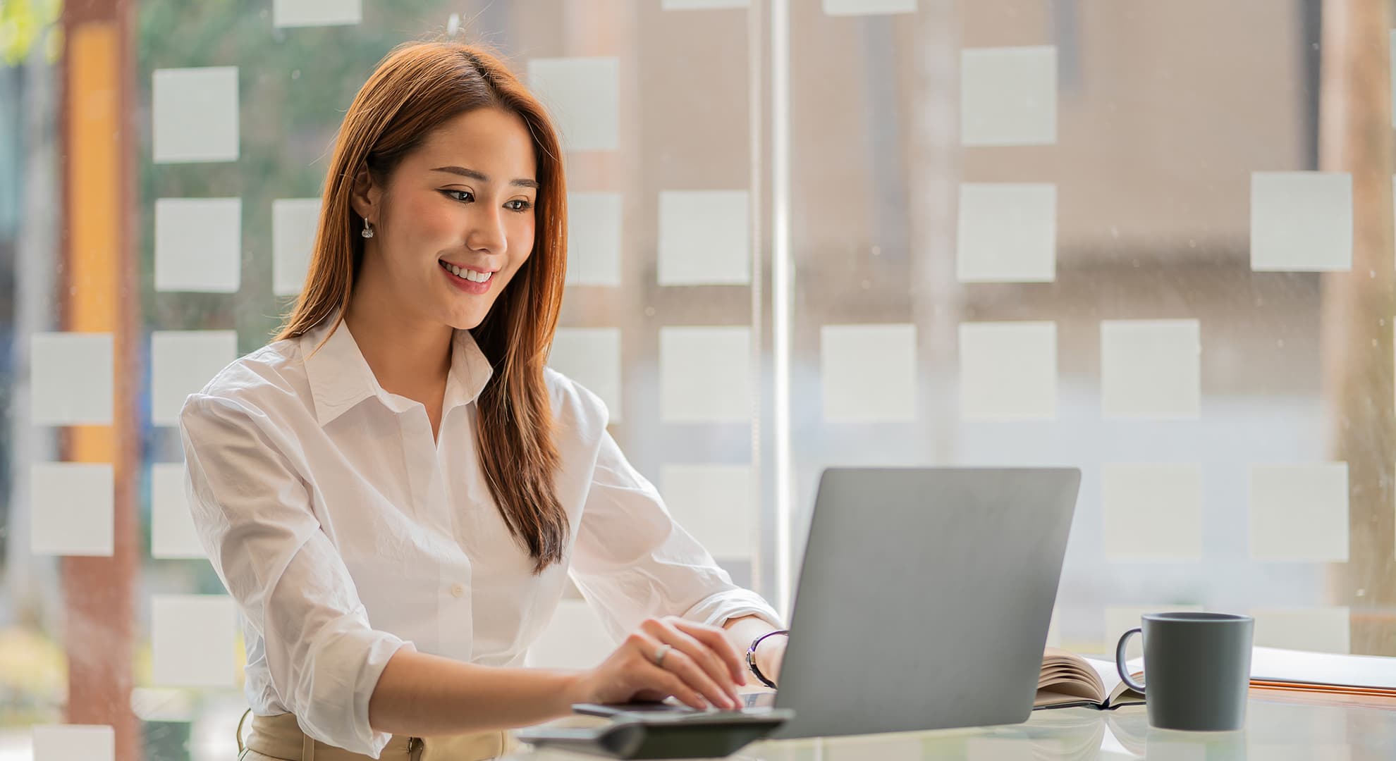 Female project manager in crisp white business shirt smiling while looking at laptop seated in office with glass behind her surrounded by planning notes.