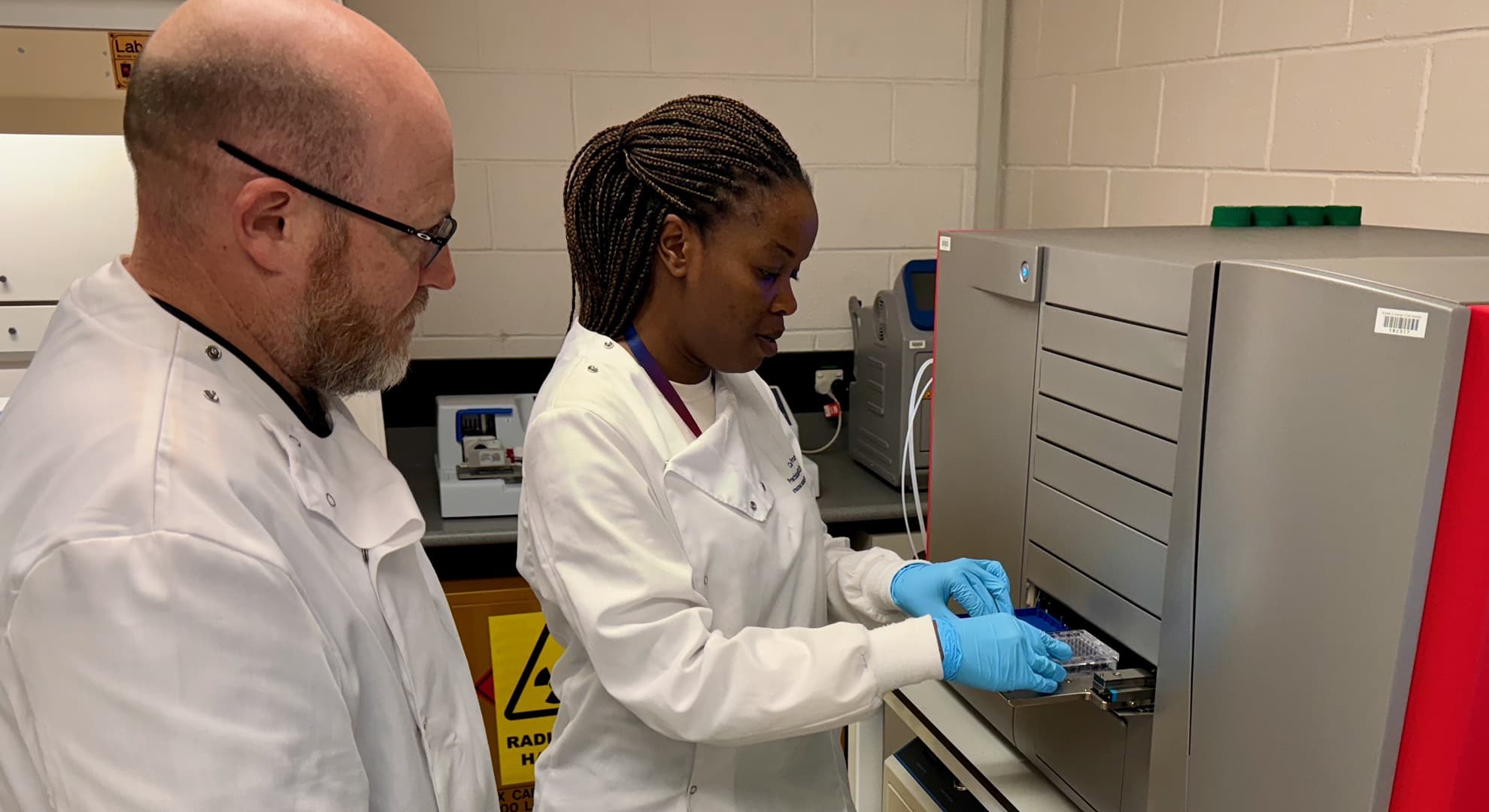 Two people in profile in white coats facing silver box-like machine, fair skinned man in foreground, dark skinned woman with blue gloves putting samples into machine's draw