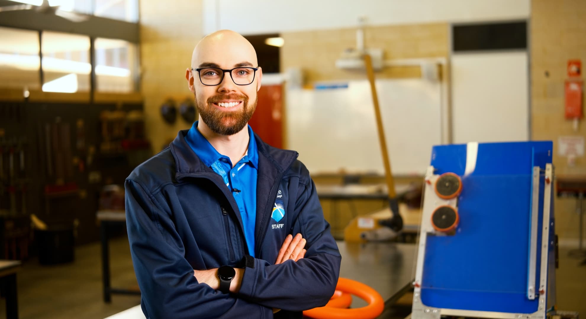 Bald young man with big smile, brown beard, glasses, navy blue zip up jacket over a bright blue polo shirt standing with arms folded in a workshop next to a bright blue cut board on an easel