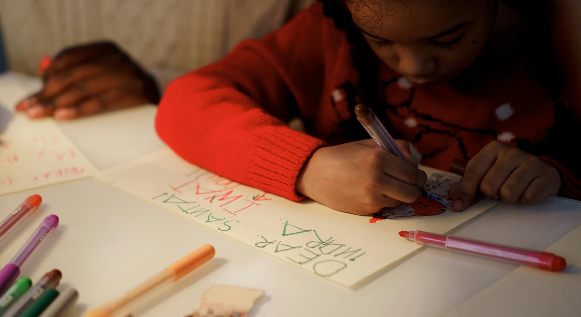 A close up overhead shot of a young girl writing a card to Santa, with the hands of her mother next to her.