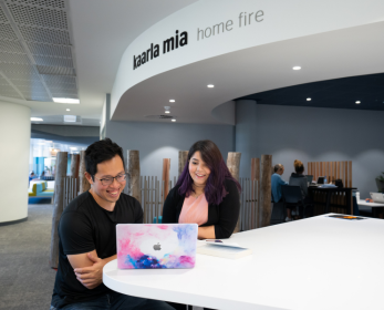 Two students sitting at a white table in ECU Library’s kaarla mia space, smiling while looking at a colourful laptop. The words 