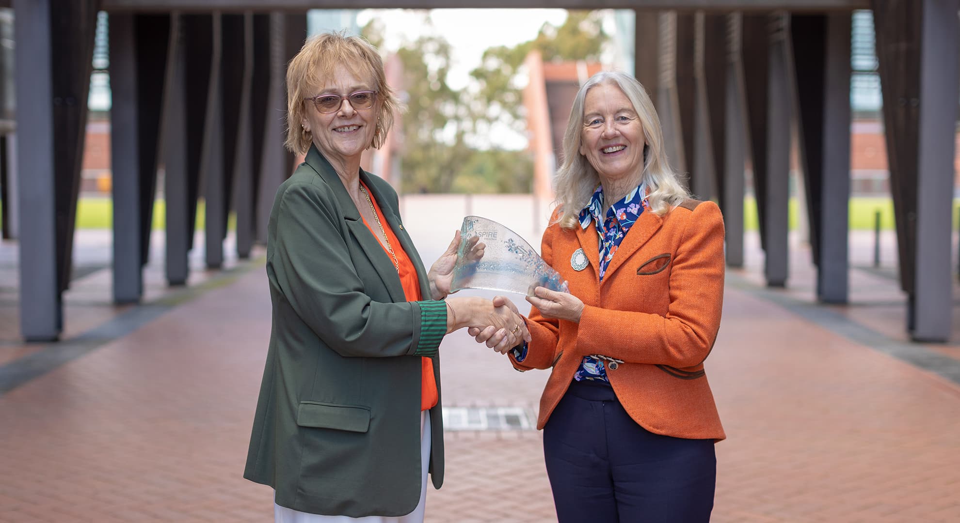 Two women outside in central walkway standing facing each other shaking  hands passing clear glass award