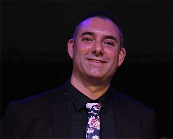 A portrait image of Mr Peter Shanhun smiling in front of a black background wearing a button down shirt and neck tie.
