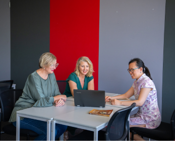 Three people sit at a table in a modern workspace, engaged in discussion. One person types on a laptop while the others look on attentively. A book and a notebook are on the table, emphasizing a focus on learning and collaboration.