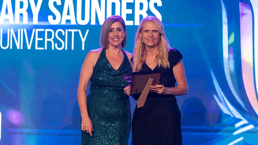 Joanne Reid wears a sparkly halter-top dress and stands with Associate Professor Rosemary Saunders who wears a black long dress and holds her framed award