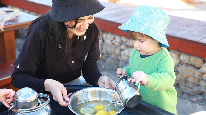 An Atlantis Group Early Childhood Educator helping a young boy pour water into a bowl.