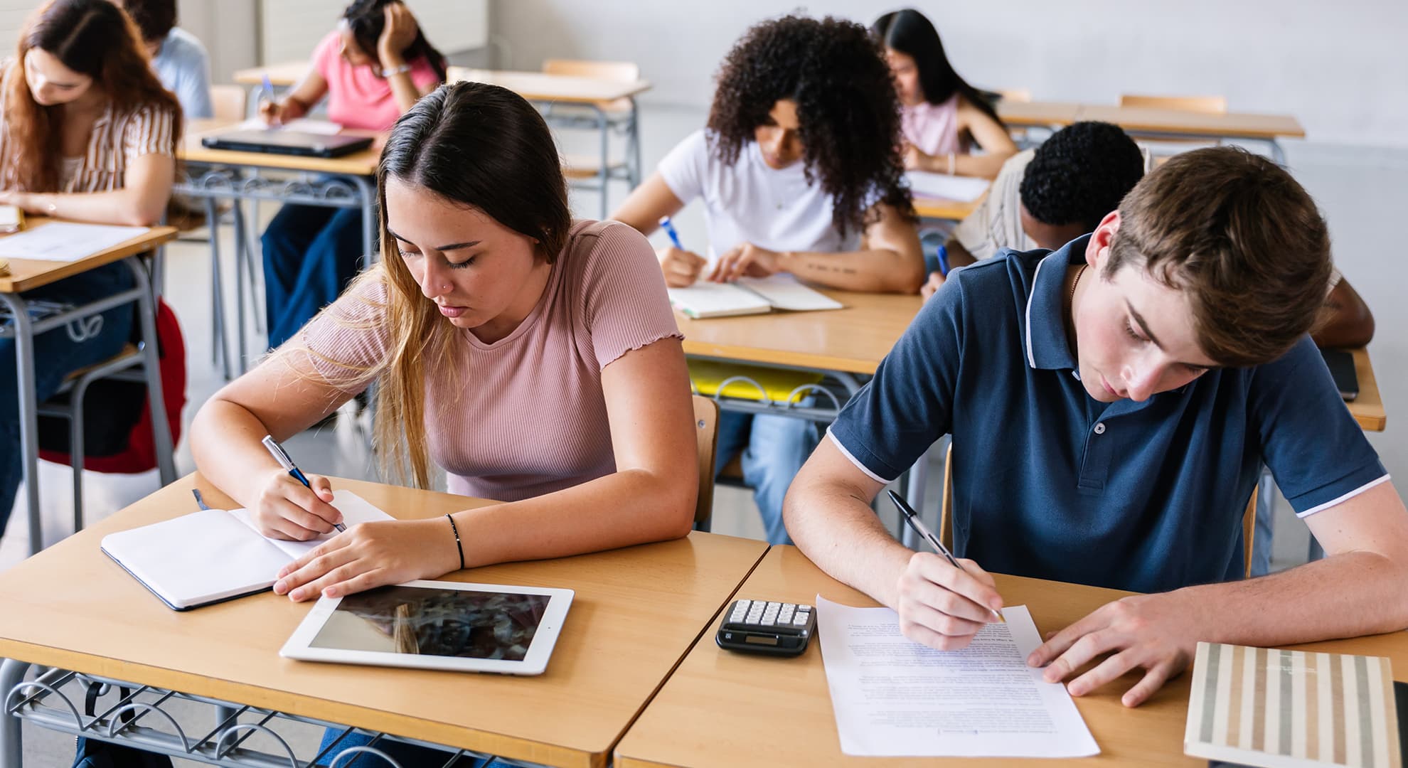 High school students sitting at school desks in a classroom, bent over their papers and devices while writing.