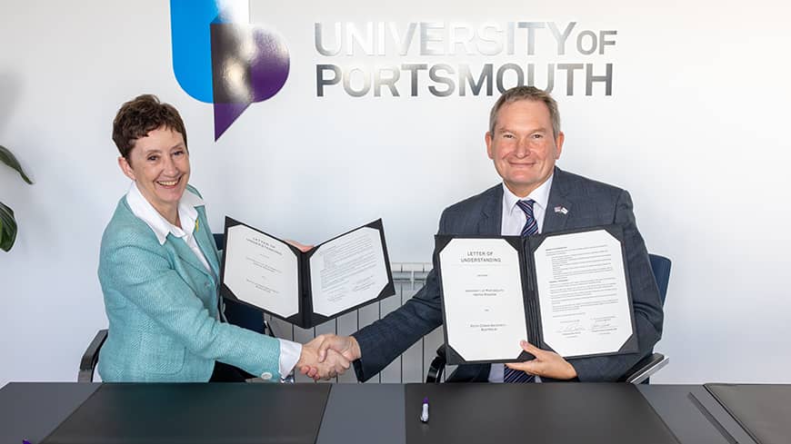 Two people sitting at a table shaking hands whilst looking at the camera, each holding a copy of a letter of understanding