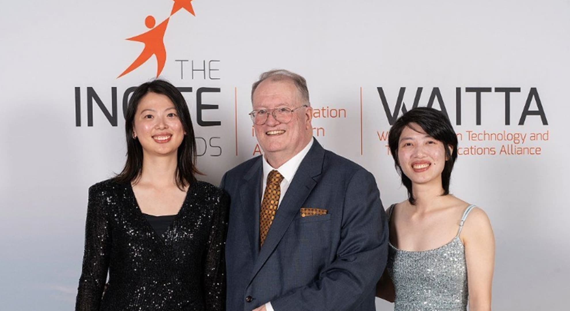 Young woman in black gown next to man with black suit next to young woman in silver gown in front of branded banner for INCITE Awards