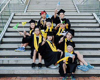 A group of Children's University students celebrate on steps in their caps and gowns waving flags.