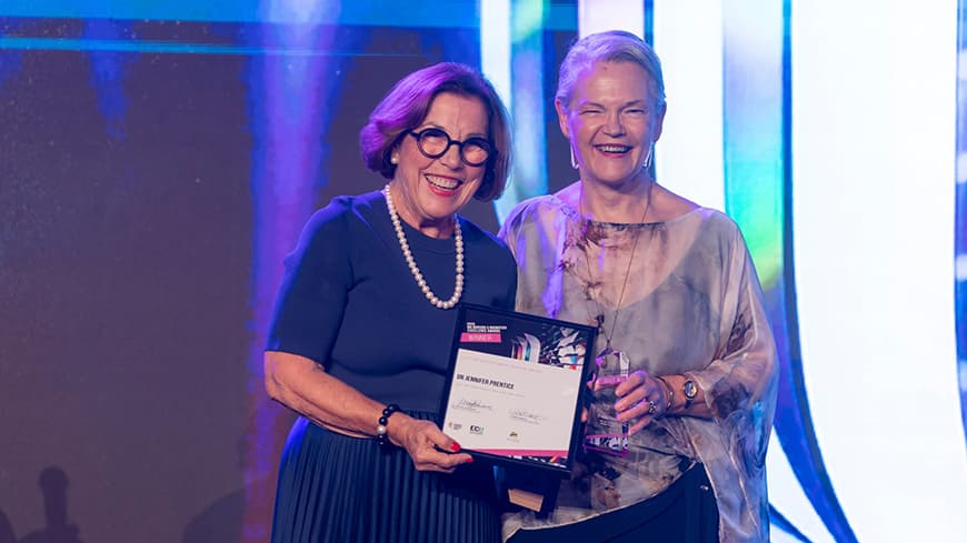 Jennifer Grieve who wears a dark blue dress, black glasses and pearls presents a framed award to Dr Jennifer Prentice who wears a flowy dress and smiles.