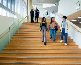 Five students walking down a wide staircase inside a modern university building, talking and smiling, with natural light and open study spaces visible.