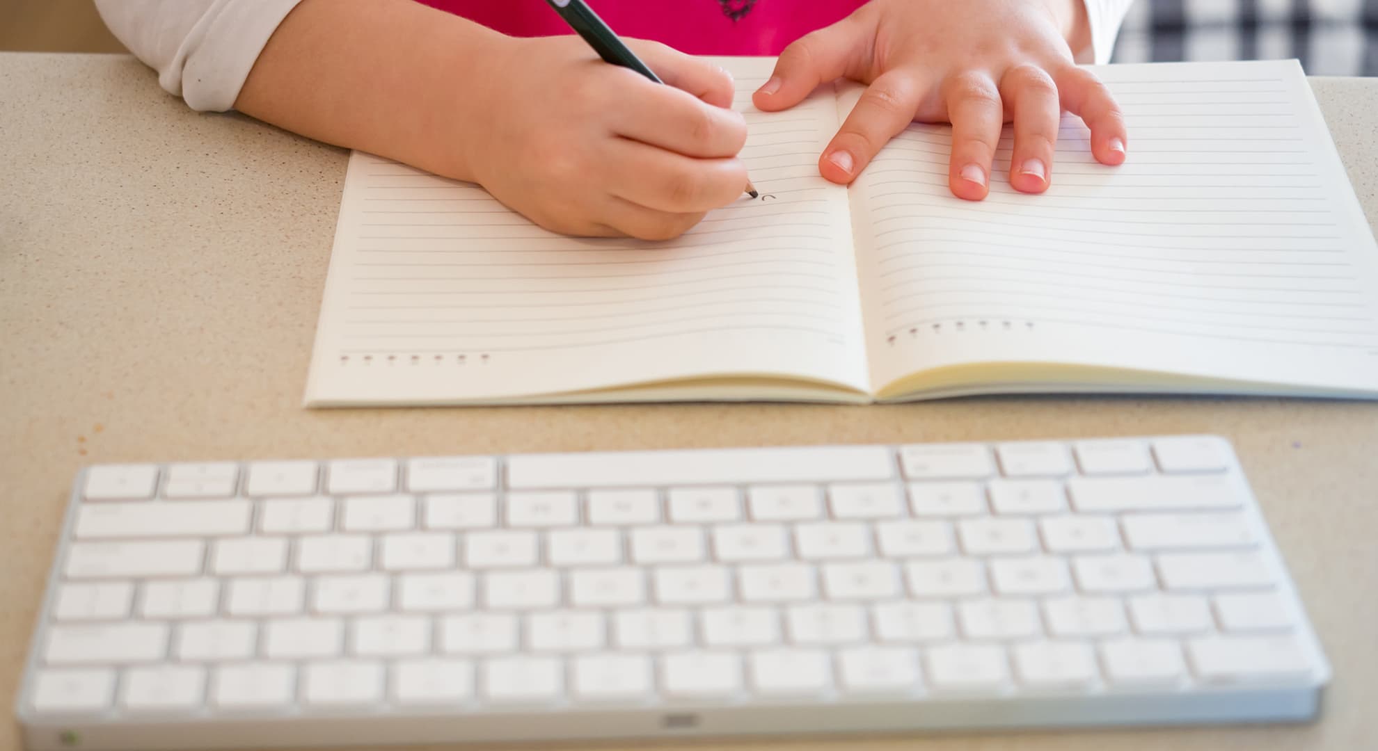 Close up shot of young child's hands holding a pen, writing on paper, with a keyboard in front of the workbook she is writing in.