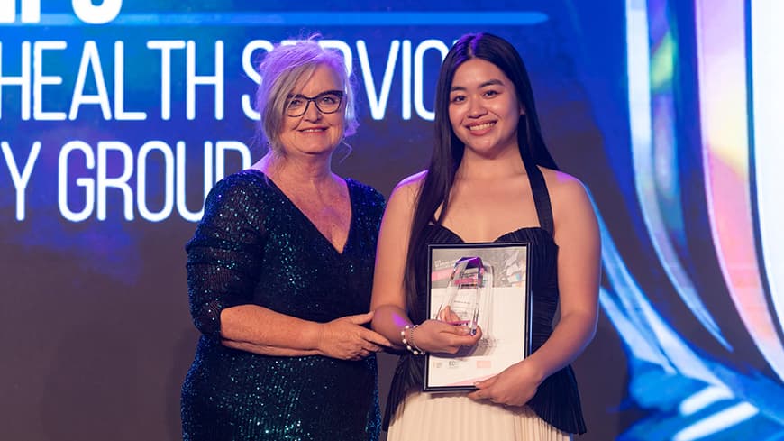 Associate Professor Vicki Cope wears a sequined dress and presents a framed award to Francine Ocampo who wears a halter-top black and white dress.