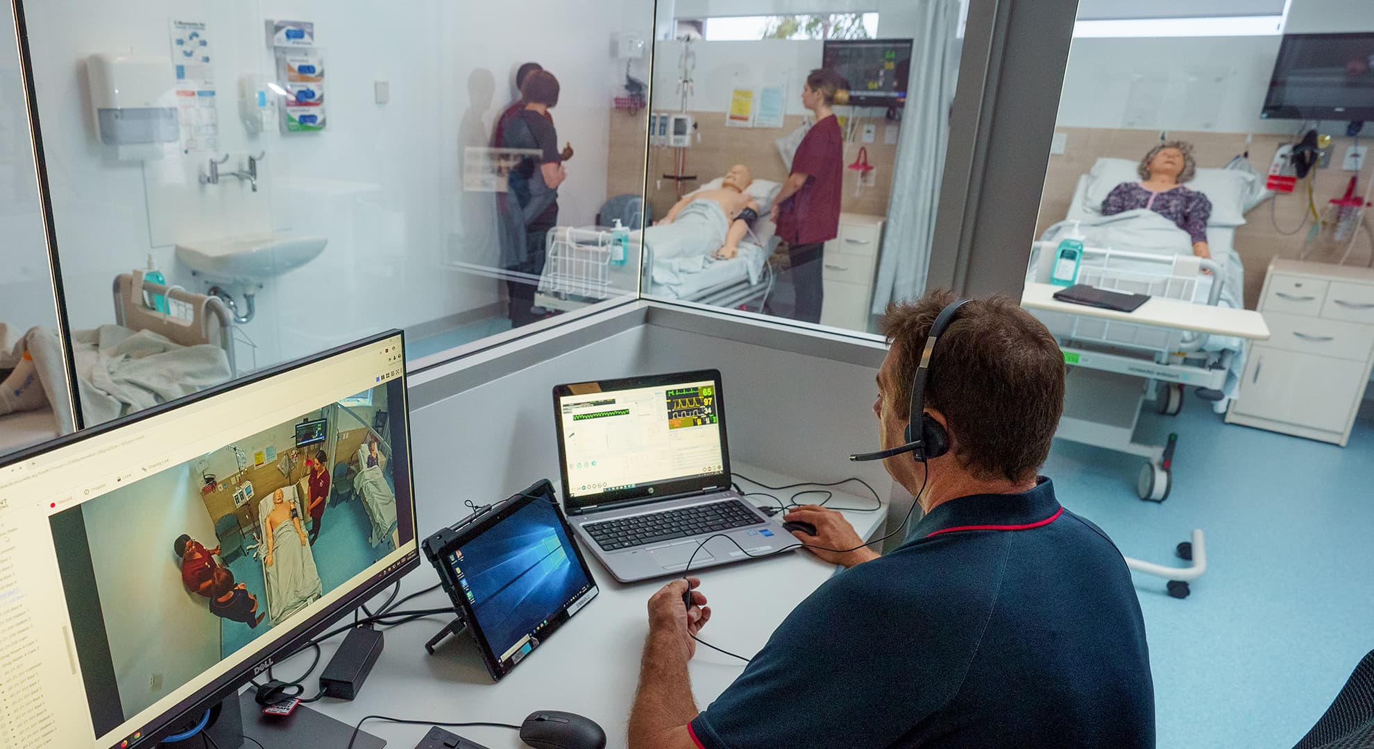 Man sitting behind glass, viewing nursing students practicing on mannequins.