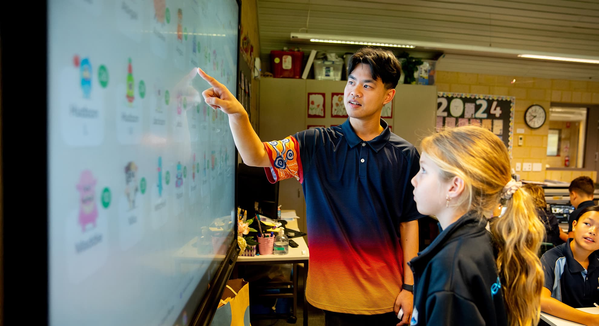 A teaching student points to the screen and explains something to a primary school student.
