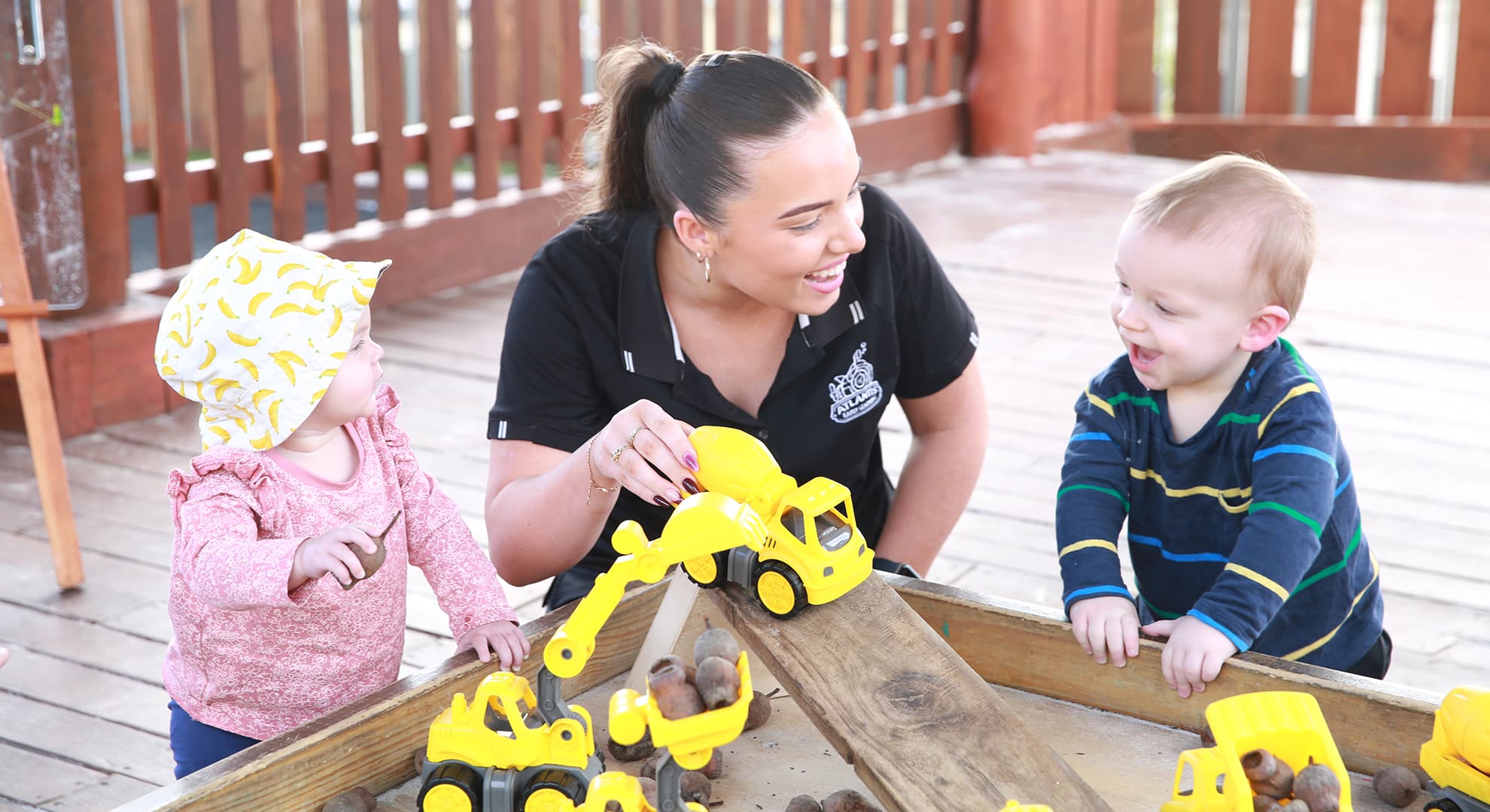 The Atlantis Group Early Childhood Educator smiling and playing trucks with two toddlers.