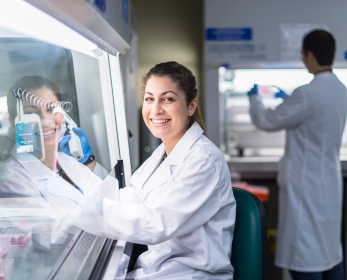 Researcher in a lab coat smiling while working in a laboratory, with another researcher blurred in the background.