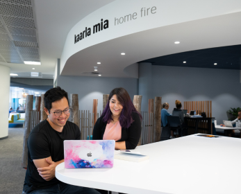 Two students sit at a white table with a laptop, smiling and working together in the 