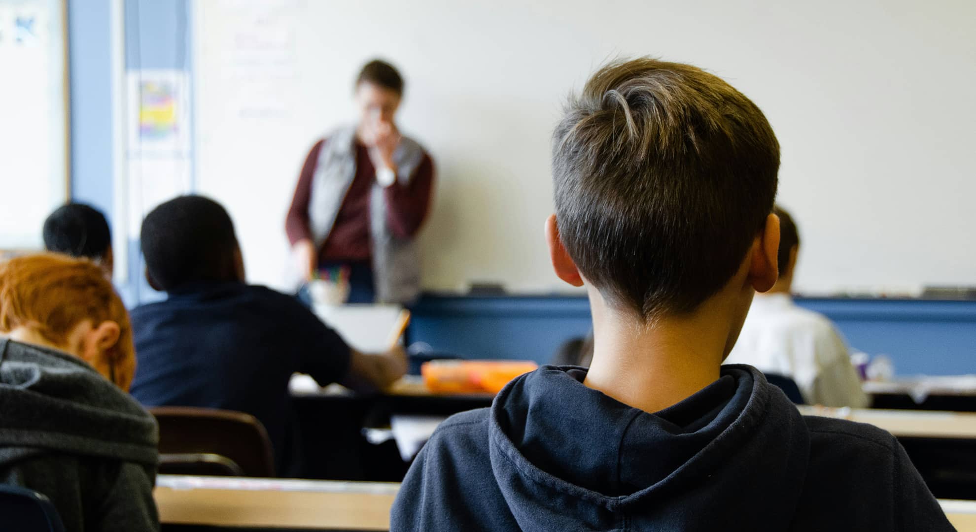 Back of high school male student\'s head in focus, looking towards teacher at the front of the class with other students and teacher out of focus in background.