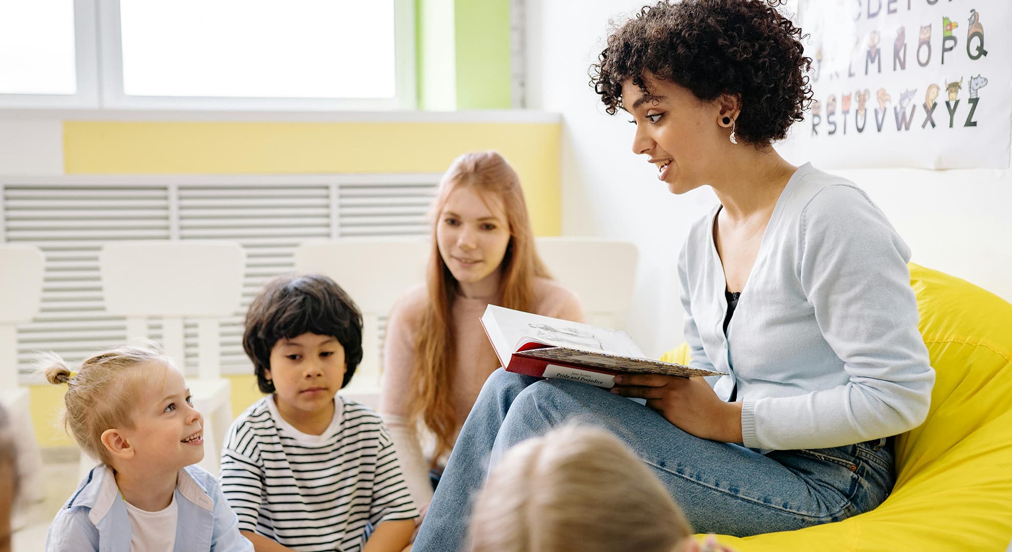 Kindy or pre-primary teacher sitting and reading to young children. Support staff are also sitting with the children.