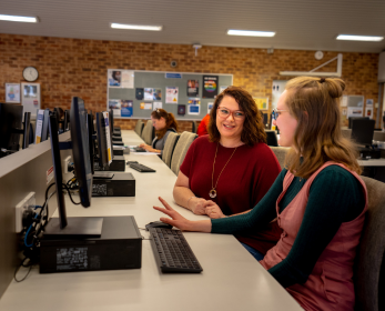 Two students seated at desktop computers in a university library, talking and collaborating, with other students working in the background.