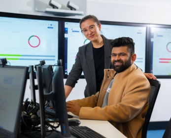 Two ECU students working at computer stations in a library technology space, with large screens displaying data visualisations in the background.