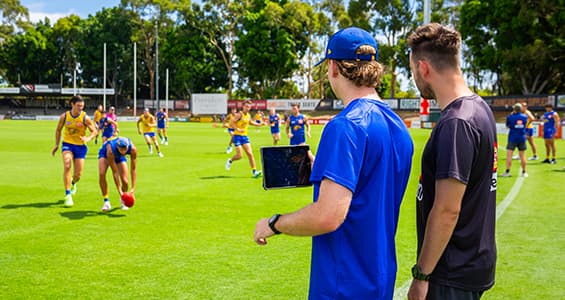 student consulting clipboard while football players run across field