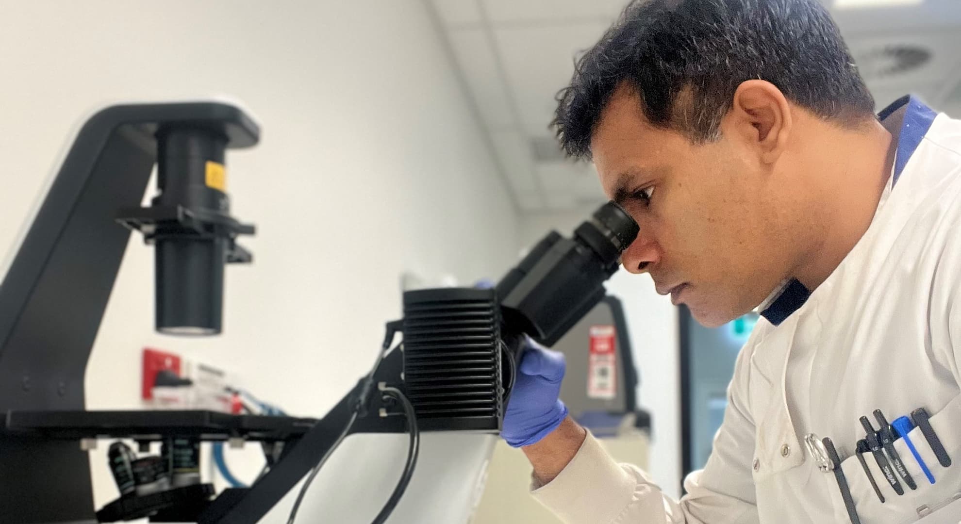 Dr Prashant Bharadwaj wears a white lab coat and blue rubber gloves, he looks into a microscope.