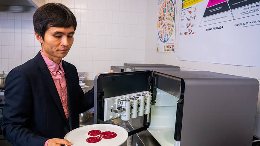 A man in front of a machine, holding a plate of red printed food.